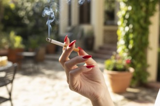 Elegant female hand with red nails holding a burning cigarette. Summer sunlight in outdoor setting