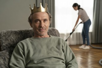 Portrait of middle-aged man with golden crown relaxing on couch while woman vacuums in background.