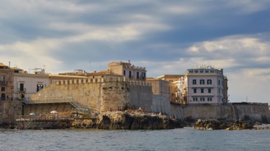 Stony coast with historic buildings under a cloudy sky, boat trip around the island of Ortigia,