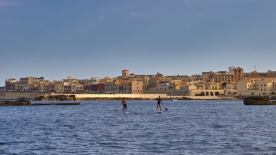 Two paddleboarders on the sea in front of a town with historical buildings under a blue sky, boat