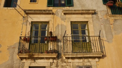 Weathered historic building façade with two balconies, windows and flowers under sunlight, Ortigia