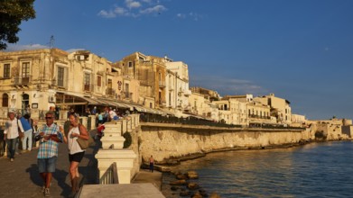 People walking along the coastal promenade with historic buildings in the evening light, Ortigia