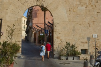 People crossing a historic stone city gate surrounded by plants, Ortigia Island, Isola di Ortigia,
