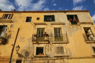 Shows the weather-beaten facade of a building with balconies and windows, blue sky above, Ortigia