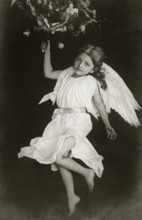 Child angel, 1930s, A child in an angel costume with wings holding a Christmas tree branch,