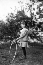 Tyre toy, 1920s, A young girl with a hula hoop stands next to a tree and looks curiously,