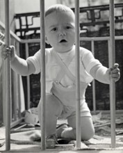 Little boy in a playpen, 1930s, Toddler kneeling in a cot and holding on to the wooden railing,