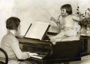 Mother with child at the piano, 1920s, A girl sits happily on a piano while a woman plays,