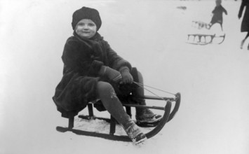 Child sledging, 1930s, A child sits on a sledge in the snow with a joyful expression on his face.