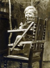 Baby in a child seat, 1930s, A smiling toddler sits happily in a wooden high chair, outside in a