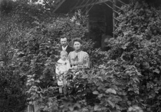 Family standing at a wooden hut, 1920s, adults with child standing in front of a house surrounded
