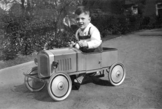 Child driving toy car, 1930s, Smiling boy driving a toy car on a street surrounded by gardens,