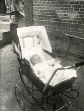 Baby in a pram, 1930s, A baby lies peacefully in a well-padded pram outside a brick building,