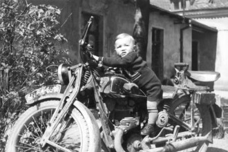 Child on a motorbike, 1930s, Small child sitting smiling on an old motorbike in a rustic setting,