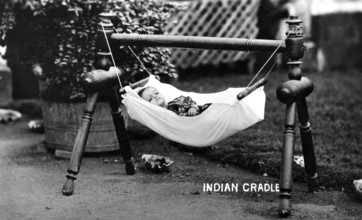 Baby hammock cradle, 1930s, A baby lies in a hanging cradle in the garden, surrounded by plants,
