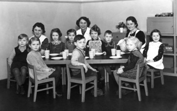 Kindergarten group, 1930s, Kindergarten scene with seated children, woman at the table with cups,