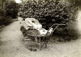 Baby in a pram, 1920, A toddler sits smiling in an antique pram in a garden, Historical photo