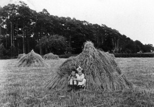2 children in a haystack, 1920, Two children sitting in a field in front of a large bale of straw,