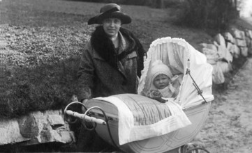 Woman with pram, 1910s, Woman with hat and coat standing with a baby in a pram on a country road,
