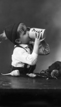 Child with a beer mug, 1930s, A boy with a cap drinks from a beer mug with 'Cheers' written on it,