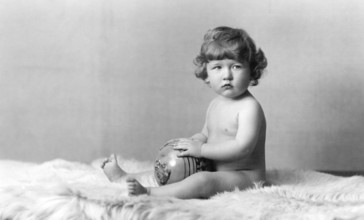 Naked child with ball, 1930s, A baby sits naked on a fur rug and holds a ball in his hands,