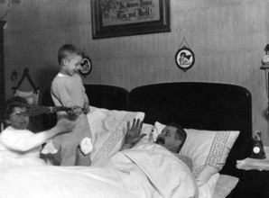 Children teasing father, 1920s, Children playing with a man in bed while one smiles and the other