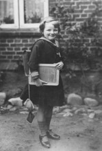 School enrolment, 1920s, A girl with a slate and school uniform stands in front of a brick wall,