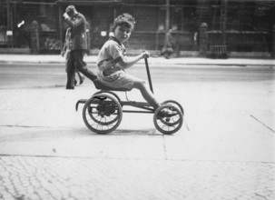 Little boy on a quadricycle, 1920s, Boy on a tricycle in an urban environment with passers-by in