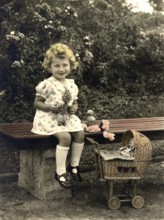 Little girl playing dolls, 1920s, Girl sitting on a bench with doll and toy trolley, surrounded by