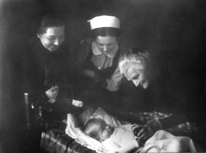 Women and baby, 20s, 1920s, Three woman gently looking at a sleeping baby in a dim light,