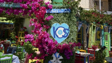 Colourfully decorated taverna with flowers and heart-shaped kos sign, sunny atmosphere, Platia
