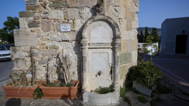 Small stone fountain by an old wall, surrounded by plants under a blue sky, Minaret, Eski Cami, Kos