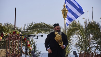 A priest in a black robe holds a golden object surrounded by palm trees in front of a Greek flag,