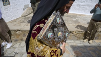 A priest carries an ornate book during a procession, dressed in richly decorated fabrics, Orthodox