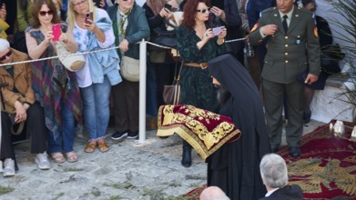 A priest in a black cloak with golden embroidery at a religious event, surrounded by people taking