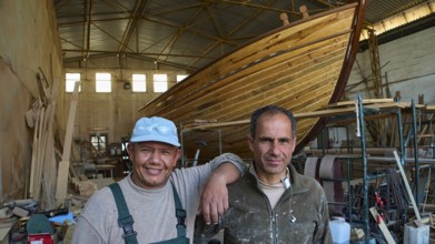 Two workers in a shipyard next to a large wooden boat hull, Diakofti boatyard, new construction of