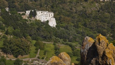 White building from a distance in a wooded landscape with rocks in the foreground, Monastery of the