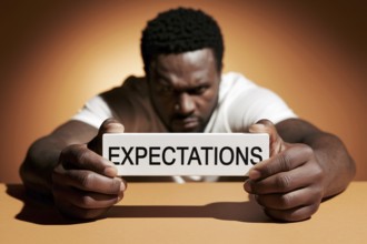 Black man holding block labeled expectations in orange studio, symbolizing social pressure and