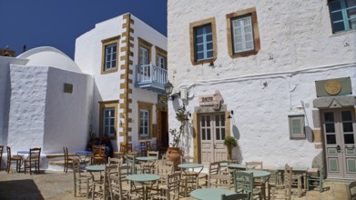 An outdoor area of a café with empty tables and chairs in front of white buildings on a sunny day,