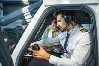 Two male pilots are sitting in the cockpit of a flight simulator, using headsets and holding