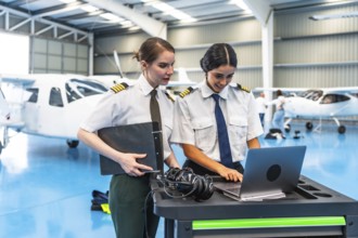 Two smiling female pilots in uniform working on a laptop inside a hangar, collaborating next to an