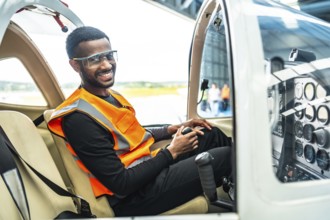 Smiling aircraft engineer sitting in cockpit holding control stick, wearing safety glasses and vest