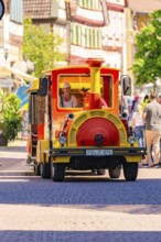 Small colourful train runs through a lively old town with cheerful people, Calw station festival,