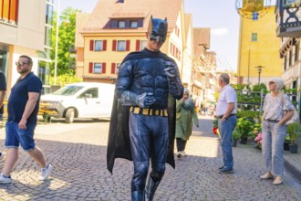 Man in Batman costume walking in a busy sunny city centre, Calw train station festival, Black