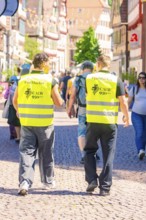 Two men in yellow waistcoats walk along a busy, sunny street, Calw railway station festival, Black