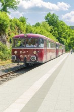 Red train stops in a rural setting on a sunny platform under a blue sky, Calw railway station