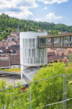 A modern building with a bridge in a town surrounded by green landscape and houses, Zob extension