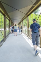 People walk over a modern glass bridge in daylight, Zob extension with bridge construction for the