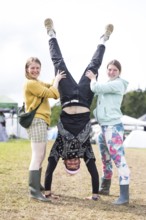 A festival visitor with a handstand thanks to the support of two woman on a camping site at the
