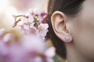 Close-up of heart-shaped pink earrings on woman's ear with cherry blossoms in warm sunlight.